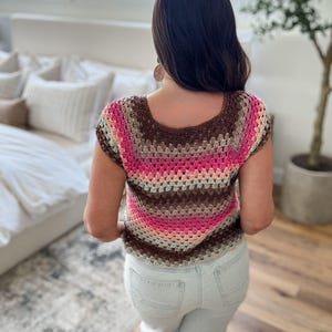 May include: A crochet top in shades of brown, pink, and beige. The short-sleeved top has a square neckline and a striped pattern. The person is wearing light blue jeans. The top is handmade.
