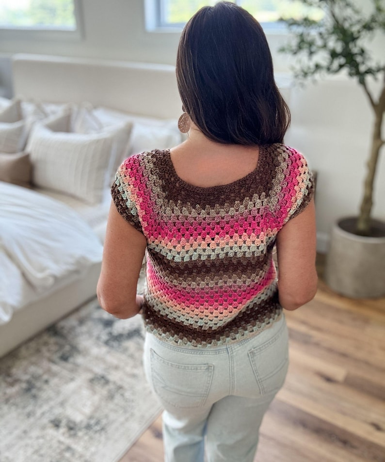 May include: A crochet top in shades of brown, pink, and beige. The short-sleeved top has a square neckline and a striped pattern. The person is wearing light blue jeans. The top is handmade.