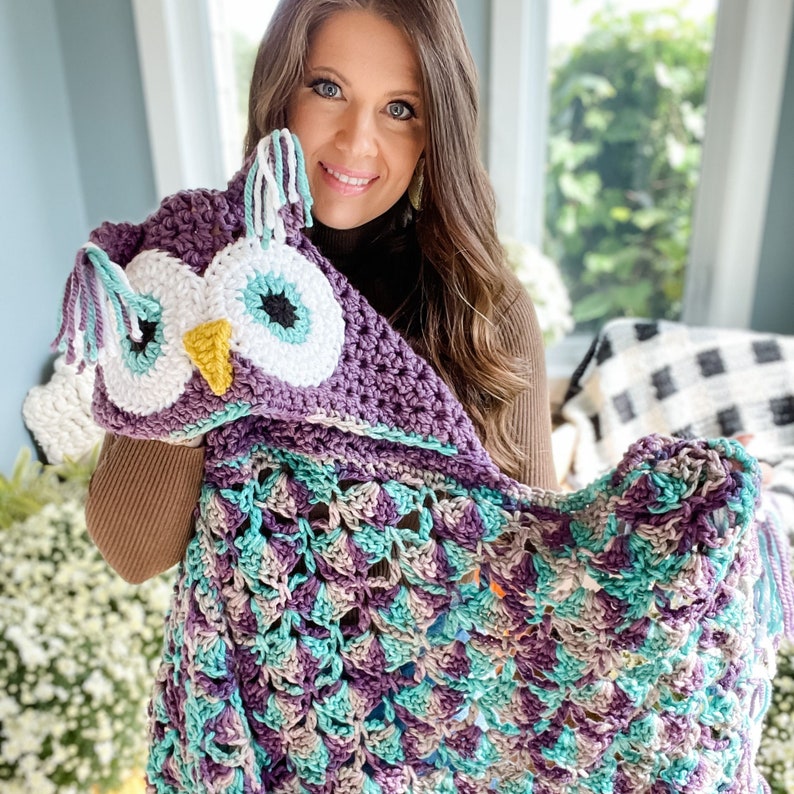 May include: A purple and teal crocheted owl hooded blanket with large white eyes and a yellow beak. The blanket is being held by a person.