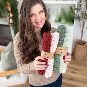 May include: A woman holds bundles of knitted washcloths in shades of red, pink, and green. Each bundle is wrapped with a brown paper band. The washcloths appear to be handmade and are displayed in a well-lit indoor setting.