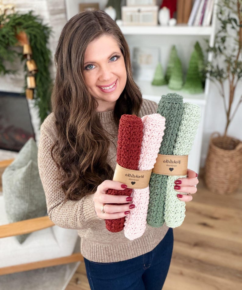 May include: A woman holds bundles of knitted washcloths in shades of red, pink, and green. Each bundle is wrapped with a brown paper band. The washcloths appear to be handmade and are displayed in a well-lit indoor setting.