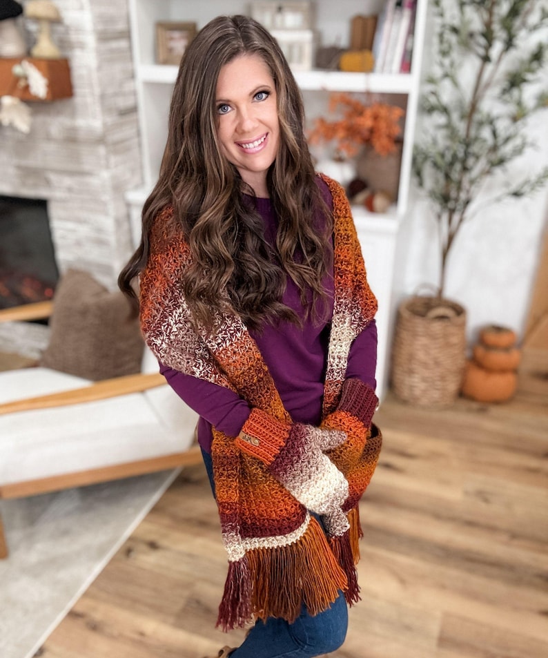 May include: A woman wearing a crocheted scarf and matching fingerless gloves. The scarf and gloves feature a gradient of colors, including orange, brown, and cream, with a fringed edge. The woman is wearing a purple long-sleeved shirt and blue jeans.