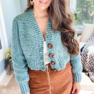 Op de afbeelding: Een tealblauwe gehaakte cardigan met bruine knopen. De cardigan is kort en heeft lange mouwen. Het model draagt een wit tanktopje onder de cardigan en een bruine corduroy rok.