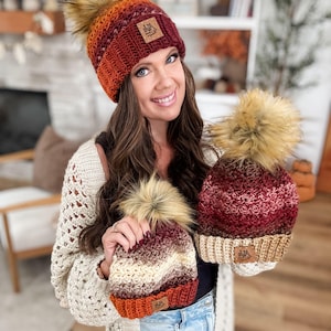 Puede incluir: Varios gorros de crochet hechos a mano en tonos rojos, naranjas y crema, cada uno rematado con un pompón de piel sintética. Los gorros tienen una etiqueta de cuero con un logotipo. La mujer lleva una rebeca de crochet color crema.