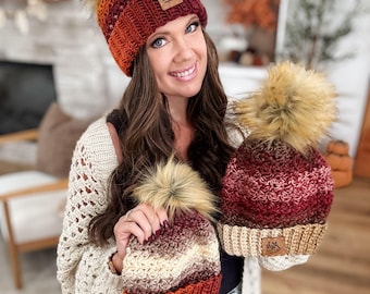 May include: A collection of handmade crochet beanies in shades of red, orange, and cream, each topped with a fluffy faux fur pom-pom. The beanies feature a leather tag with a logo. The woman is wearing a cream-colored crochet cardigan.