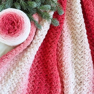 May include: A soft, textured blanket in shades of red, pink, and cream. A ball of yarn with similar color gradients sits next to the blanket. Green pine branches are placed on top of the blanket and yarn.