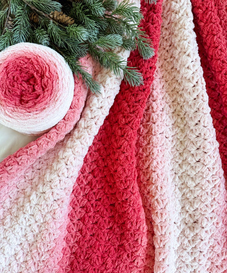 May include: A soft, textured blanket in shades of red, pink, and cream. A ball of yarn with similar color gradients sits next to the blanket. Green pine branches are placed on top of the blanket and yarn.