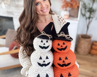 Puede incluir: Dos pilas de calabazas de ganchillo, una blanca y otra naranja, cada una con una cara sonriente de jack-o'-lantern. Las calabazas están rematadas con sombreros de bruja negros, una decoración festiva de Halloween.