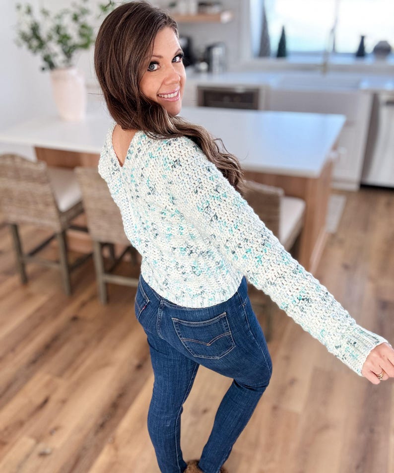 May include: A white and blue speckled sweater with a V-neck and long sleeves. The sweater is worn over blue jeans.