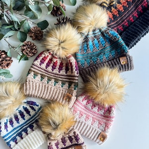May include: Six crocheted winter hats with faux fur pom poms. The hats feature a geometric pattern in various colours, including teal, pink, purple, and brown. Each hat has a brown leather tag with the word "Mila" on it.