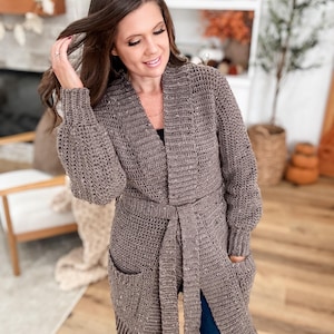 Può includere: Cardigan lungo marrone all'uncinetto con cintura e tasche. Il cardigan ha maniche lunghe e un motivo a punto strutturato. La cintura si stringe in vita. Il cardigan è indossato sopra un top nero e jeans blu.