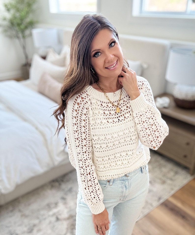 K&ouml;nnte beinhalten: Ein cremefarbener H&auml;kelpullover mit langen &Auml;rmeln und rundem Ausschnitt. Der Pullover hat ein offenes, spitzenartiges Design. Die Person tr&auml;gt eine goldene Halskette und hellblaue Jeans. Der Hintergrund umfasst ein Bett und einen Nachttisch.