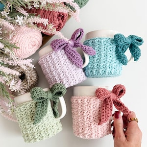 May include: Four white ceramic mugs with crocheted cozies in pastel colors: lavender, teal, sage green, and rose. Each cozy has a matching bow. The mugs are arranged on a white surface, with yarn and flowers in the background.