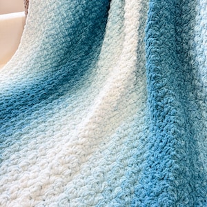 May include: A soft, textured blanket in shades of blue and white. The blanket features a gradient effect, transitioning from a deep blue to a light, airy white. The fabric has a raised, textured pattern, adding visual interest and a cozy feel.