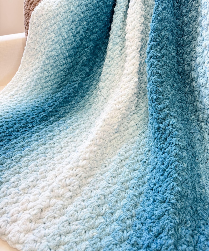 May include: A soft, textured blanket in shades of blue and white. The blanket features a gradient effect, transitioning from a deep blue to a light, airy white. The fabric has a raised, textured pattern, adding visual interest and a cozy feel.