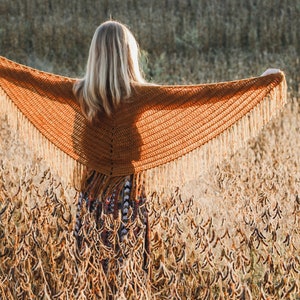 Puede incluir: Una mujer con un chal de ganchillo marrón con flecos, de pie en un campo de plantas secas.