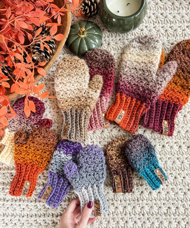 Peut inclure: Une collection de mitaines crochet&eacute;es &agrave; la main, aux couleurs vari&eacute;es : beige, orange, violet et turquoise. Chaque mitaine pr&eacute;sente un poignet c&ocirc;tel&eacute; et une petite &eacute;tiquette en cuir. Les mitaines sont dispos&eacute;es sur une surface textur&eacute;e de couleur cr&egrave;me.
