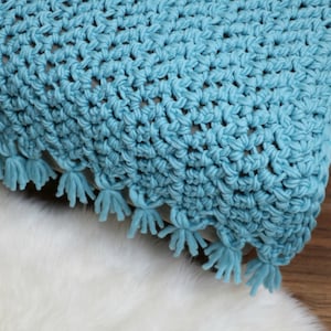Pu&ograve; includere: Una coperta di uncinetto blu con un bordo a frangia. La coperta &egrave; realizzata con un semplice motivo a punto e ha una consistenza morbida.