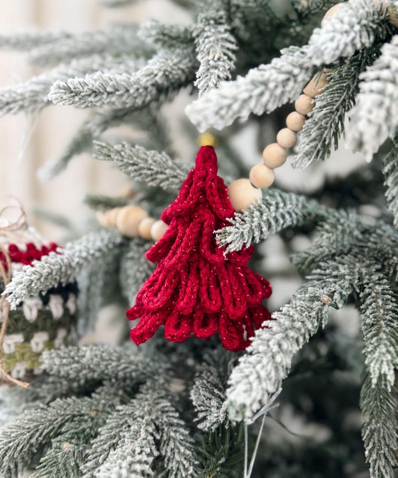 K&ouml;nnte beinhalten: Nahaufnahme eines roten, geh&auml;kelten Weihnachtsbaumschmucks mit Goldglitzer, der an einem schneebedeckten Tannenzweig h&auml;ngt. Eine Holzperlen-Girlande und andere Ornamente sind ebenfalls zu sehen, was eine festliche Urlaubsszene schafft.