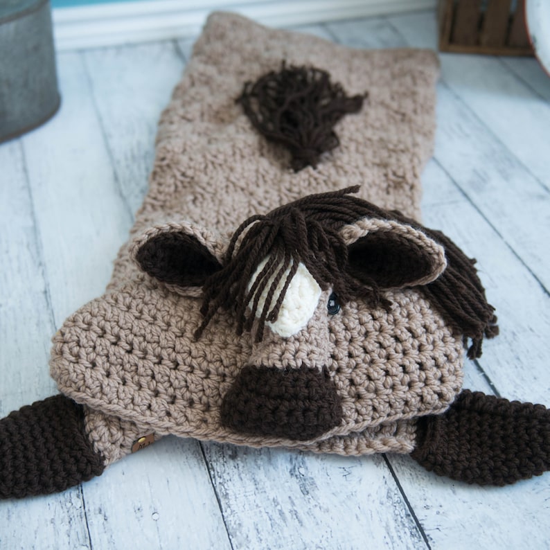 Pu&ograve; includere: Una coperta per cavallo in crochet marrone con un viso marrone e bianco, orecchie marroni e una coda marrone. La coperta &egrave; appoggiata su una superficie di legno bianco.