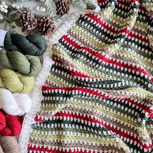 Puede incluir: Una manta de crochet a rayas en tonos de verde, rojo, blanco y marrón. La manta está hecha con un punto texturizado y tiene un borde de crochet.