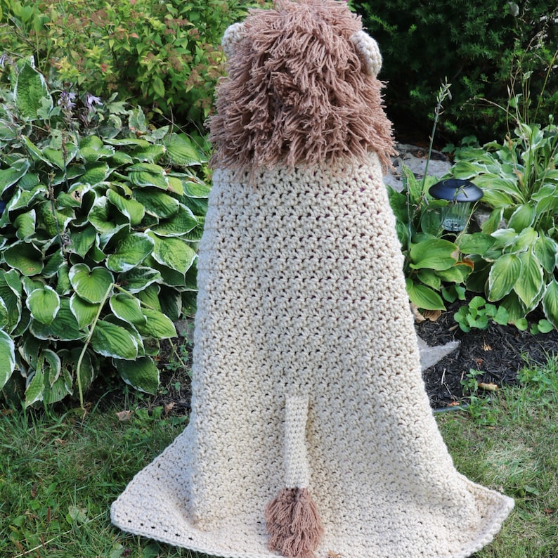 May include: A crocheted lion blanket with a brown and white color scheme. The blanket has a lion head hood and a tail with a tassel.