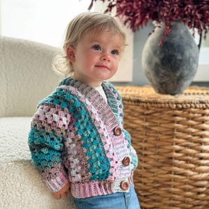 Può includere: Un bambino indossa un cardigan all'uncinetto multicolore con bottoni in legno. Il cardigan ha una combinazione di colori rosa, verde e marrone. Il bambino indossa jeans blu.