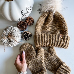 May include: A brown crocheted beanie with a fluffy white pom pom and a brown leather label that says "Mils". The beanie is paired with a matching pair of brown crocheted mittens.