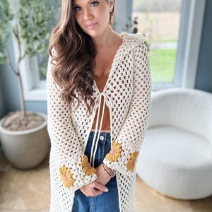 May include: A white crochet cardigan with a tie front and sunflower details on the sleeves. The cardigan is open and worn over a brown tank top and blue denim skirt.