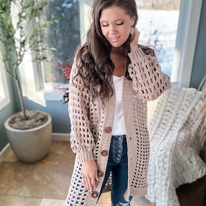 May include: A long, open-weave, beige crochet cardigan with large wooden buttons. The cardigan has long sleeves and is worn over a white top and blue jeans. The background shows a bright, sunlit room with a large window.