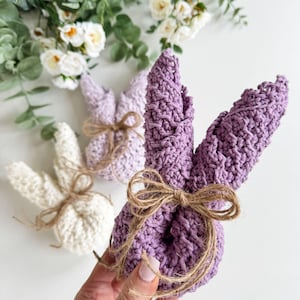 May include: Three crocheted bunny shapes, one white and two purple, tied with twine bows. The purple bunnies are held in a hand.