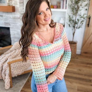 Könnte beinhalten: Ein regenbogenfarbenes Häkeltop mit V-Ausschnitt und langen Ärmeln. Das Top hat horizontale Streifen in Pastellfarben wie Rosa, Gelb, Blau und Grün. Das offene Webdesign verleiht dem Kleidungsstück eine zarte Textur. Die Person trägt blaue Jeans.