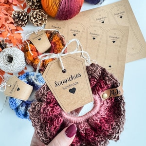 May include: Handmade scrunchies in shades of blue, orange, and burgundy, with matching tags. The scrunchies are made of yarn and have a tag that says "Scrunchies Handmade". There are also spools of yarn and pinecones.