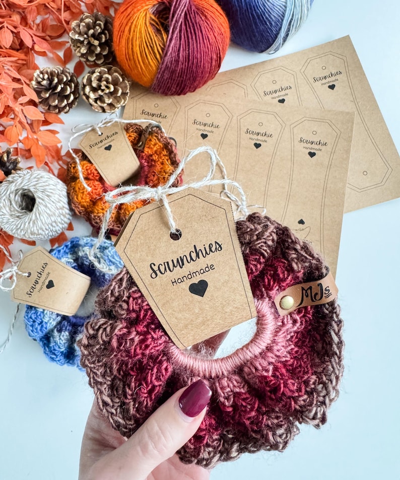 May include: Handmade scrunchies in shades of blue, orange, and burgundy, with matching tags. The scrunchies are made of yarn and have a tag that says "Scrunchies Handmade". There are also spools of yarn and pinecones.