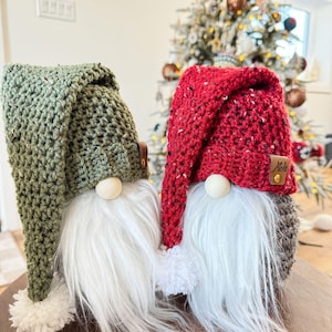 May include: Two crocheted gnomes, one with a green hat and the other with a red hat. Both gnomes have white beards and pom-poms on their hats.