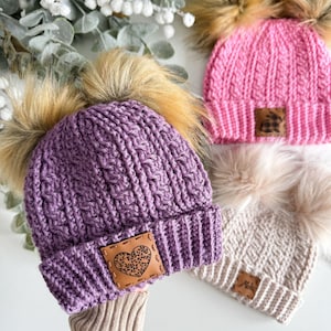May include: Three crocheted hats with faux fur pom poms. The hats are purple, pink, and beige. Each hat has a leather patch with a heart design.