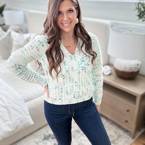 May include: A woman wearing a white and blue speckled sweater with a v-neck and long sleeves. She is also wearing dark blue jeans.