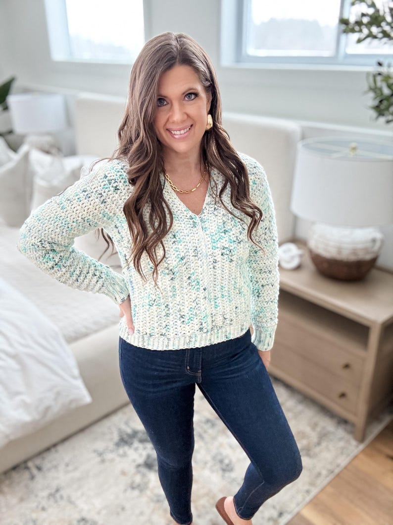 May include: A woman wearing a white and blue speckled sweater with a v-neck and long sleeves. She is also wearing dark blue jeans.