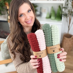May include: A woman holds two bundles of crocheted washcloths. One bundle includes a deep red and a light pink cloth, and the other includes two shades of green. Each bundle is wrapped with a brown paper band with a heart logo.