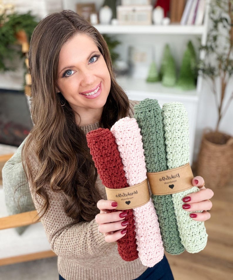 May include: A woman holds two bundles of crocheted washcloths. One bundle includes a deep red and a light pink cloth, and the other includes two shades of green. Each bundle is wrapped with a brown paper band with a heart logo.