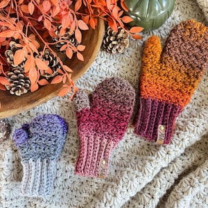 Peut inclure: Trois mitaines crochet&eacute;es dans les tons de violet, rose et orange. Les mitaines ont un poignet c&ocirc;tel&eacute; et un motif de point textur&eacute;. Chaque mitaine a une petite &eacute;tiquette en cuir. Les mitaines sont dispos&eacute;es sur une surface textur&eacute;e de couleur cr&egrave;me.