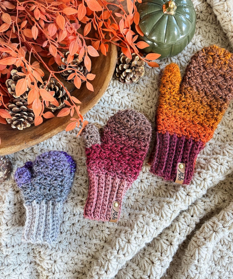 Peut inclure: Trois mitaines crochet&eacute;es dans les tons de violet, rose et orange. Les mitaines ont un poignet c&ocirc;tel&eacute; et un motif de point textur&eacute;. Chaque mitaine a une petite &eacute;tiquette en cuir. Les mitaines sont dispos&eacute;es sur une surface textur&eacute;e de couleur cr&egrave;me.