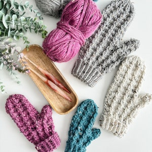 May include: Five crocheted mittens in various colors, including teal, pink, and gray, with two knitting needles and yarn balls in the background.