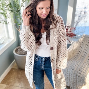 May include: A cream-colored, open-weave crochet cardigan with large wooden buttons and pockets. The long-sleeved sweater has a relaxed fit and is worn over a white top and blue jeans. The cardigan is a cozy, handmade garment.