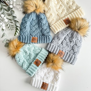 Może przedstawiać: Pięć czapek na szydełku z pomponami z imitacji futra. Czapki są w różnych kolorach, w tym białym, jasnoniebieskim, szarym i zielonym. Każda czapka ma brązową skórzaną naszywkę z tekstem "Mel's All Designs".