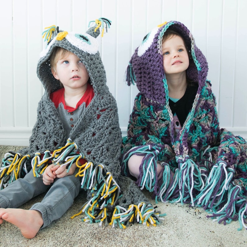 May include: Two children wearing crocheted owl hooded blankets. The blanket on the left is gray with yellow, white, and black fringe. The blanket on the right is purple, teal, and white with white fringe.