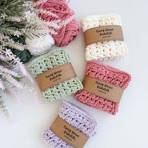 Puede incluir: Un conjunto de pa&ntilde;os de cocina Honey Weave hechos a mano en crema, rosa, verde y lavanda. Cada pa&ntilde;o est&aacute; envuelto con una banda de papel marr&oacute;n que dice "Honey Weave Dishcloth Handmade". Tambi&eacute;n hay ovillos de lana en colores a juego y acentos florales.