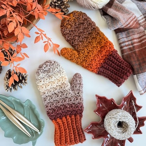 Peut inclure: Deux mitaines crochet&eacute;es &agrave; la main, dans un d&eacute;grad&eacute; de couleurs allant du cr&egrave;me au marron et &agrave; l'orange. Les mitaines sont pr&eacute;sent&eacute;es avec de la laine, des aiguilles &agrave; tricoter et des d&eacute;corations sur le th&egrave;me de l'automne.