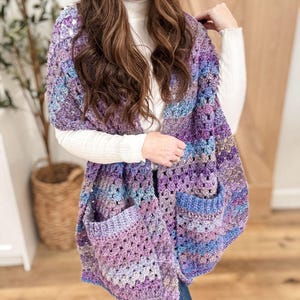 Peut inclure: Un châle en crochet violet, bleu et marron avec une grande poche. Le châle est réalisé avec un point de granny square et présente un motif rayé.