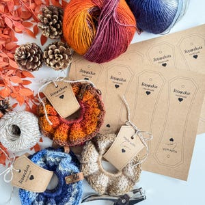 May include: An overhead shot features handmade scrunchies in shades of orange, blue, and beige, alongside skeins of yarn in complementary colors. The scrunchies have tags that read "Scrunchies Handmade" and "Made with Love". Pinecones and fall foliage add decorative elements.